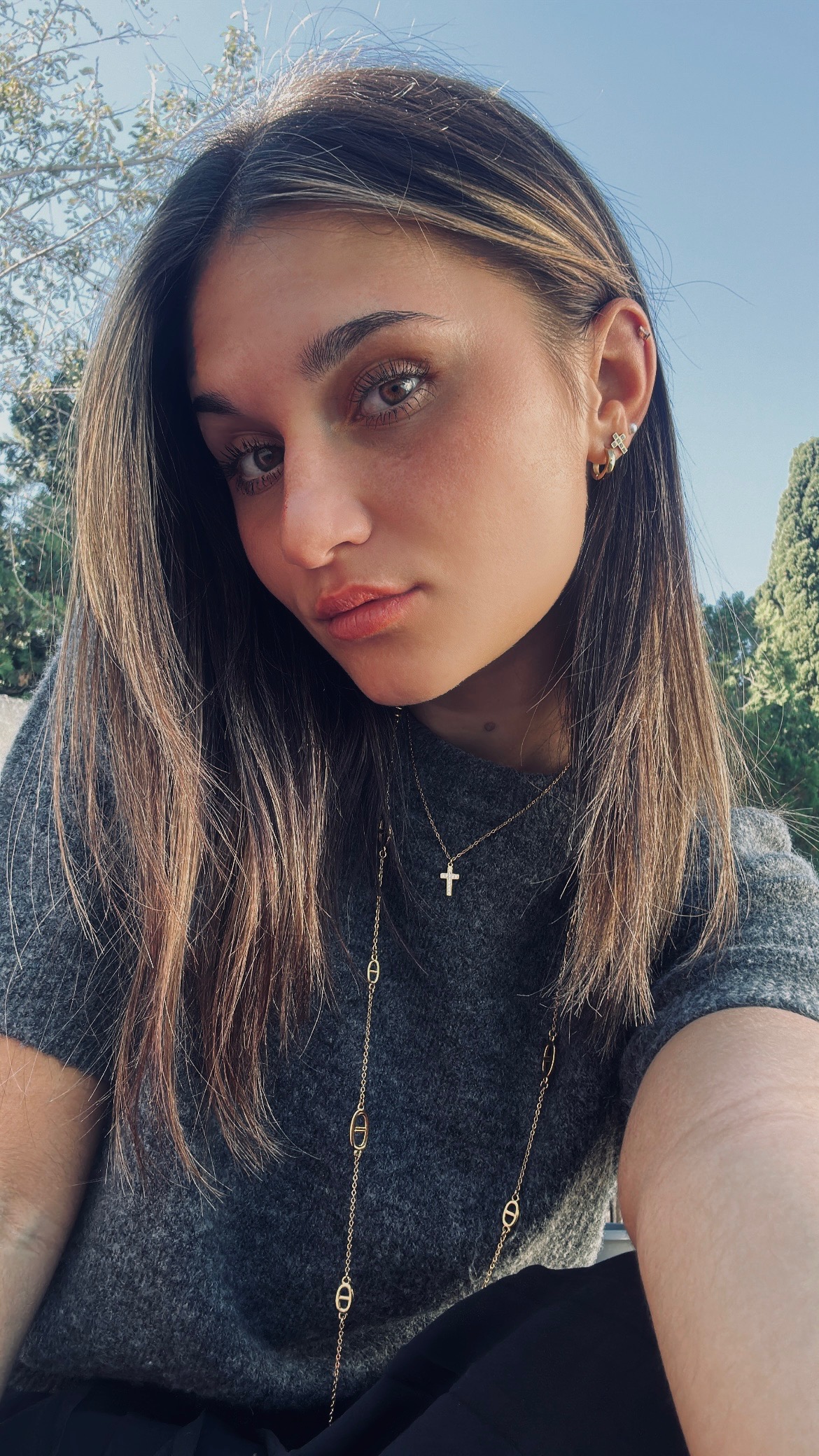 Close-up portrait of a young woman outdoors in a grey sweater, with layered gold necklaces and small earrings, against a blue sky.